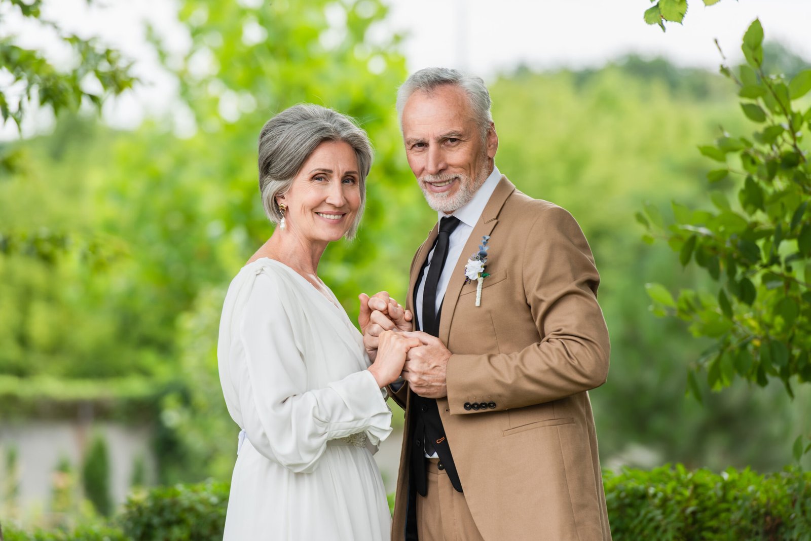 happy middle aged groom in suit holding hands with mature bride in white dress in garden